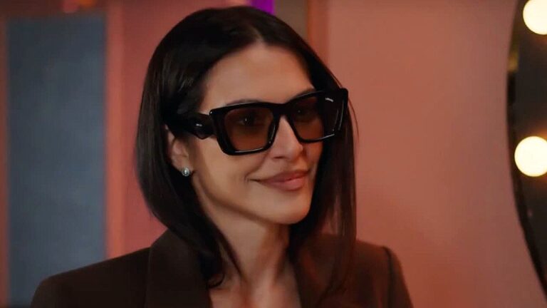 Cleo Pires com óculos escuros e blazer marrom em cena de produção audiovisual ambientada em camarim iluminado