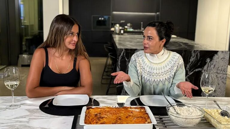 Duas mulheres conversam à mesa de jantar com travessa de lasanha em cozinha moderna durante vídeo para redes sociais