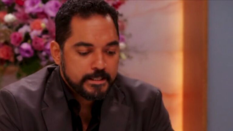 Homem de barba e paletó escuro olha para baixo com expressão séria durante conversa em ambiente elegante decorado com flores ao fundo