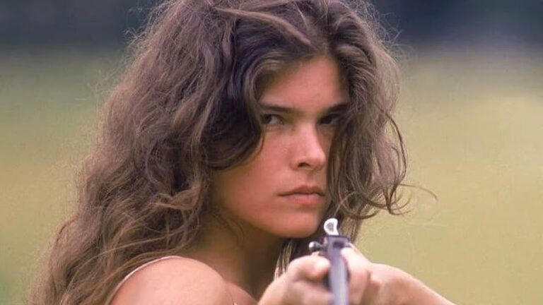 Personagem de cabelos longos e expressão determinada aponta arma em cena ambientada ao ar livre na novela Pantanal