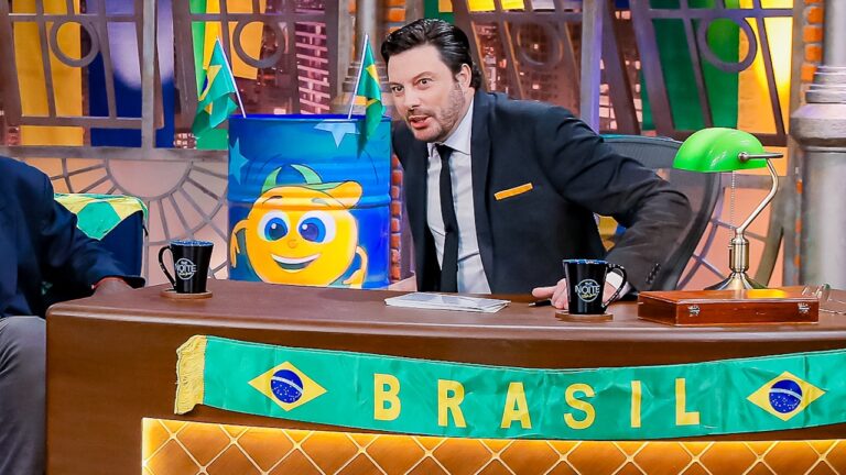Danilo Gentili, de terno escuro, inclinado atrás da bancada do The Noite com canecas do programa, bandeiras do Brasil e decoração temática ao fundo