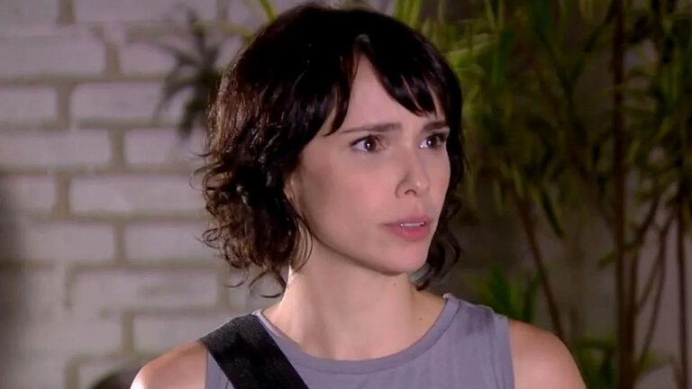 Mulher de cabelos curtos e escuros olha para o lado com expressão apreensiva em cena de novela diante de parede clara e plantas ao fundo