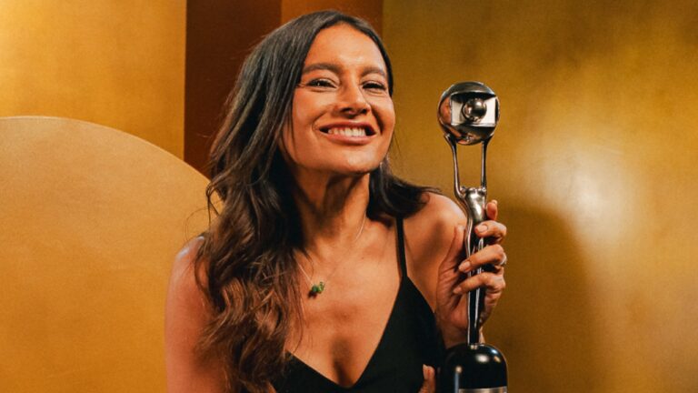 Dira Paes sorri segurando troféu prateado com vestido preto diante de fundo dourado em cerimônia de premiação