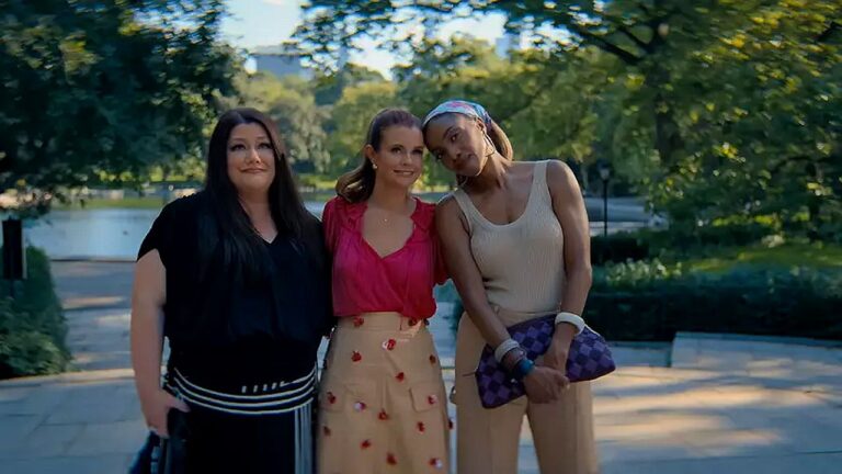 Três mulheres posam juntas em parque arborizado durante passeio ao ar livre em cena de série de comédia dramática