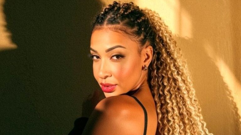 Emelly Souza com cabelo cacheado preso em tranças, maquiagem destacada e vestido preto posa de perfil diante de parede iluminada por luz dourada