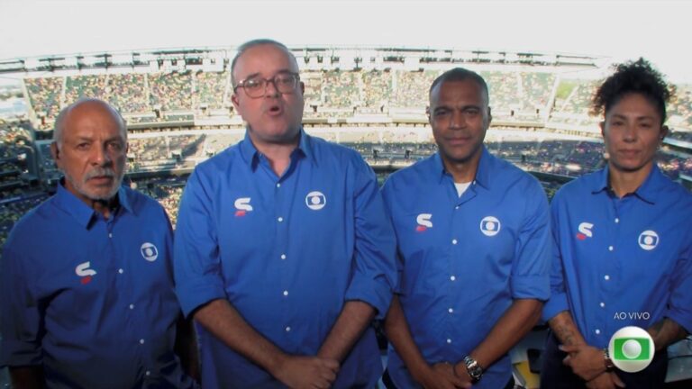 Everaldo Marques e equipe da Globo, de camisas azuis, na cabine de transmissão de Brasil x Haiti com estádio lotado ao fundo