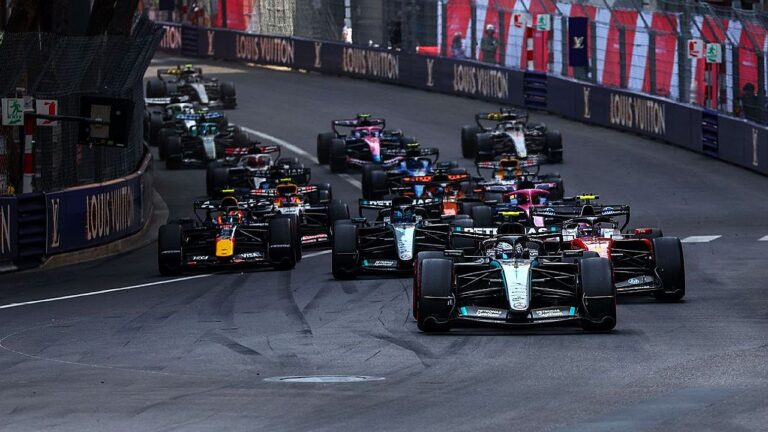 Pelotão de carros de Fórmula 1 disputa posição em curva de circuito urbano cercado por muros de proteção durante corrida automobilística internacional