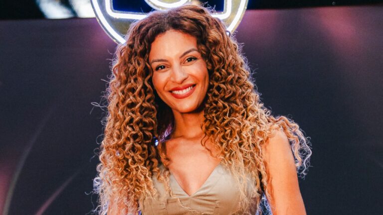 Gabriela Loran sorri para foto em evento com iluminação de palco e cabelos cacheados volumosos ao destaque