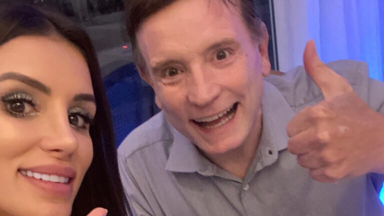 Gaby Cabrini sorri para selfie ao lado de Roberto Cabrini, que faz sinal de positivo com o polegar em ambiente interno iluminado por luz azul