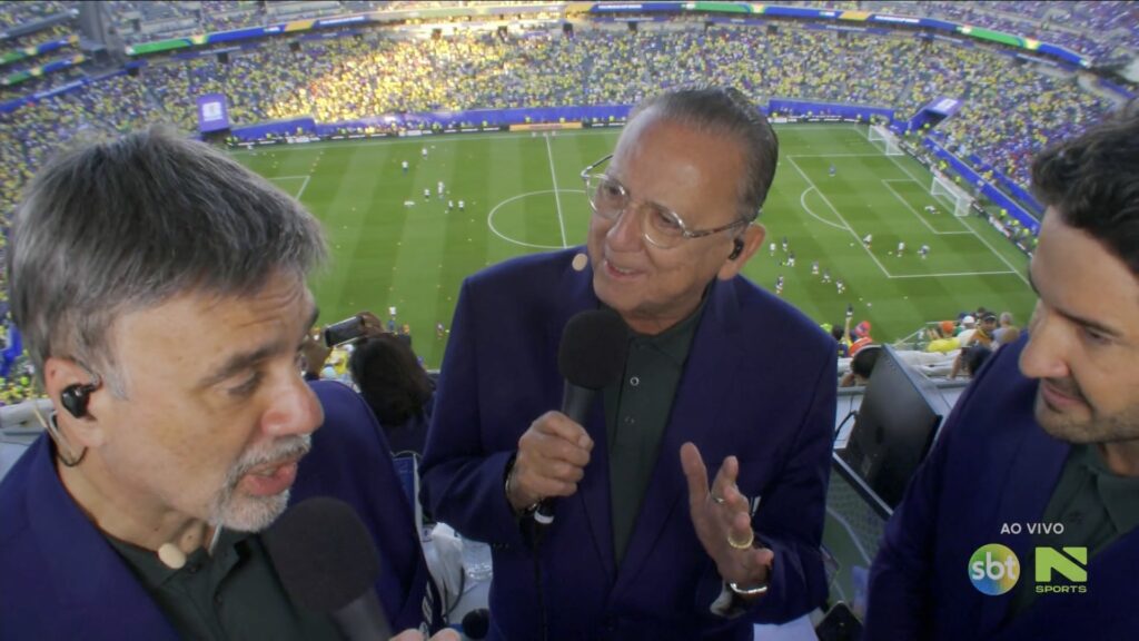 Galvão Bueno segura microfone ao lado de comentaristas na cabine de transmissão com estádio lotado e gramado ao fundo