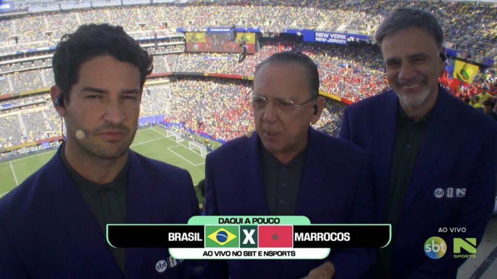Galvão Bueno entre colegas de blazer azul na cabine de transmissão com telão e torcida ao fundo