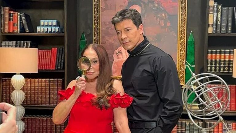 Susana Vieira segura lupa ao lado de Rodrigo Faro em cenário de biblioteca durante gravação de programa de TV