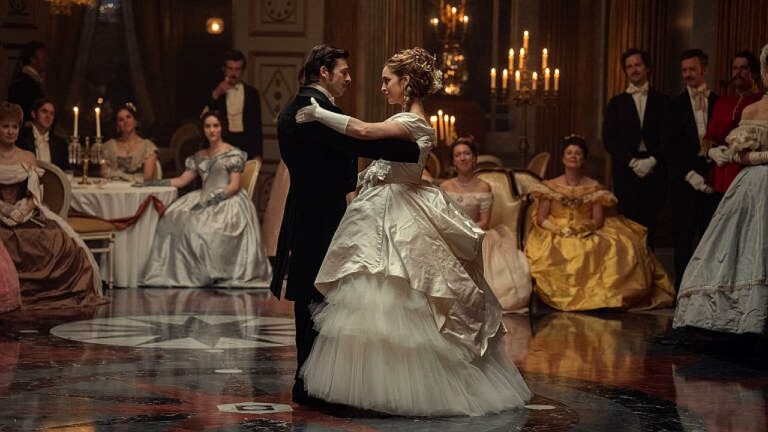 Casal dança em salão iluminado por velas durante baile da alta sociedade em cena de drama de época