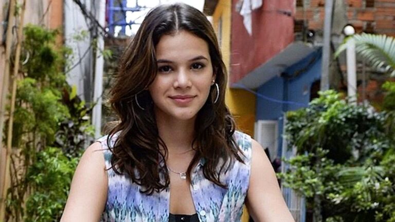 Atriz Bruna Marquezine sorrindo em rua com vegetação ao fundo em cena da novela I Love Paraisópolis