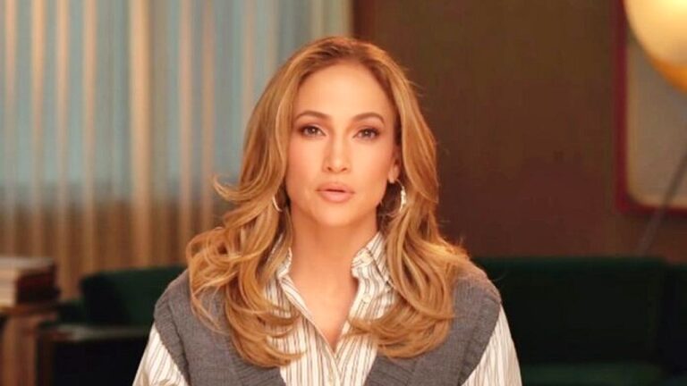 Jennifer Lopez encara a câmera durante gravação em ambiente interno usando camisa listrada, colete cinza e brincos de argola grandes