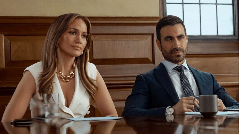 Jennifer Lopez e Brett Goldstein sentados em sala de reuniões em cena da comédia romântica Office Romance