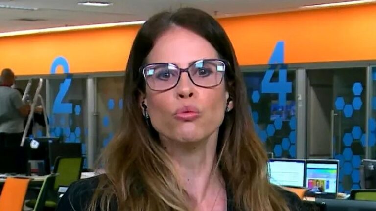 Jornalista de óculos fala ao vivo em redação de telejornal com computadores e estações de trabalho ao fundo