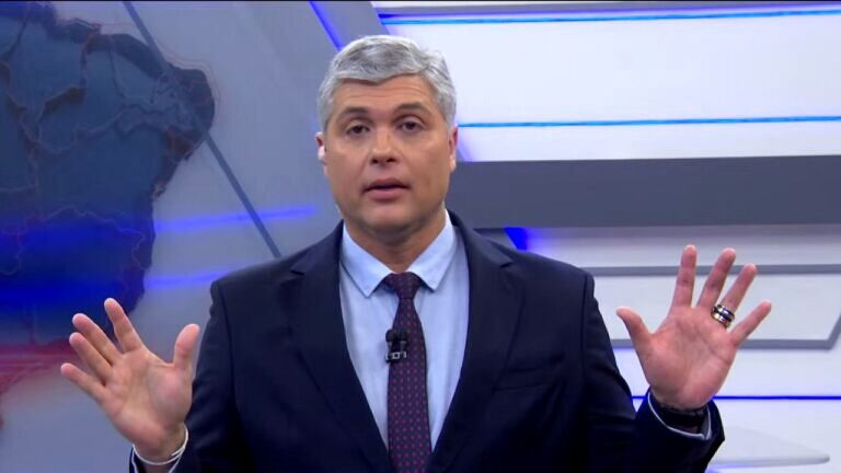 Joel Datena apresenta telejornal ao vivo com as mãos erguidas diante de painel gráfico em estúdio da Band