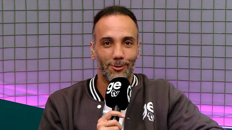 Apresentador segura microfone da ge tv durante participação em programa esportivo com cenário iluminado ao fundo