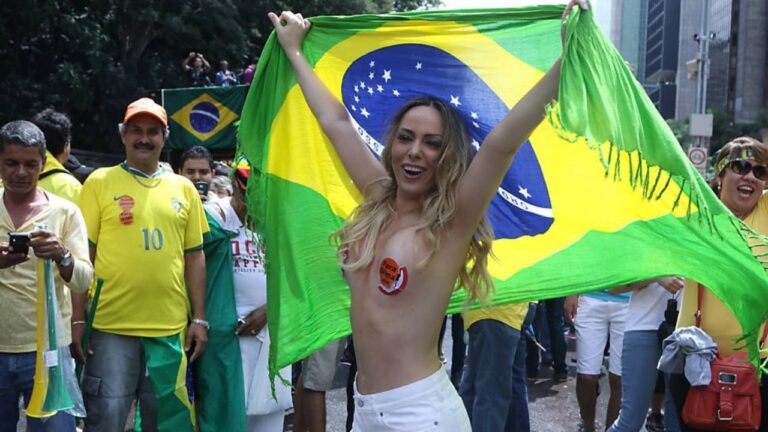 Ju Isen ergue bandeira do Brasil durante manifestação de rua cercada por apoiadores com camisetas verde e amarelas