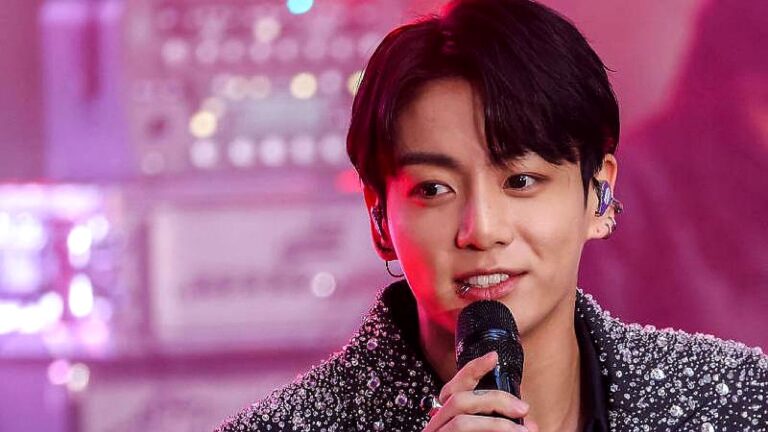 Jungkook canta ao microfone durante apresentação com figurino brilhante e iluminação colorida ao fundo