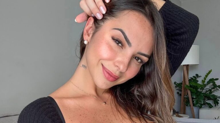 Karol Rosalin sorri para a câmera com mão na cabeça e blusa preta em ambiente interno residencial