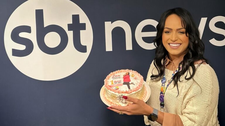Lisa Gomes segura bolo comemorativo em frente ao logotipo do SBT News durante celebração de um ano na emissora