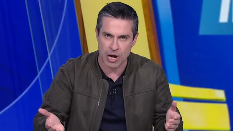Luciano Amaral de jaqueta verde gesticula durante apresentação em estúdio com painel azul e amarelo ao fundo