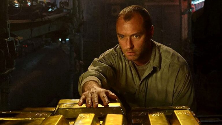 Jude Law observa barras de ouro em ambiente industrial em cena do thriller de ação Mar Negro