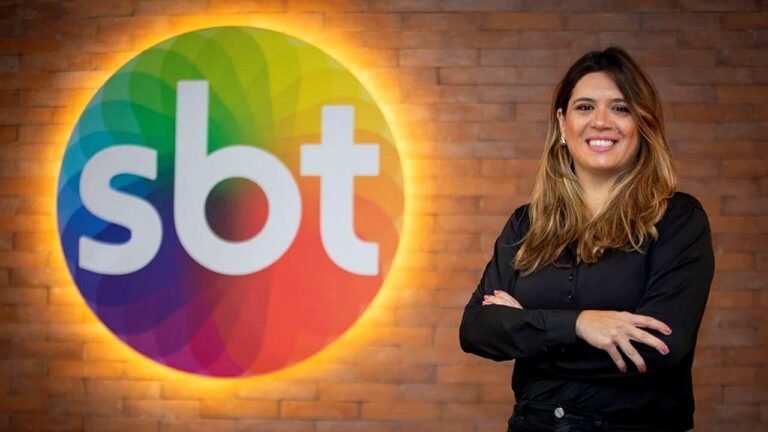 Executiva do SBT posa sorrindo diante do logotipo da emissora em escritório com parede de tijolos ao fundo