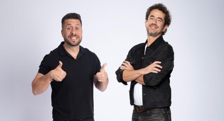 Dois apresentadores posam para foto promocional com roupas escuras e gestos descontraídos diante de fundo neutro