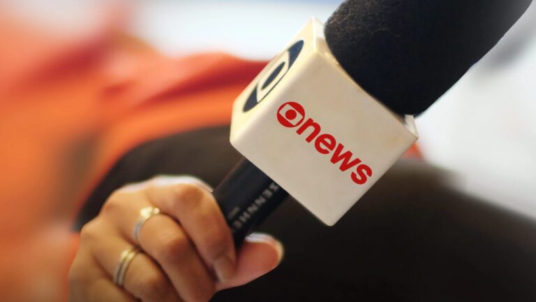 Microfone com logotipo da GloboNews segurado por repórter em ambiente desfocado ao fundo