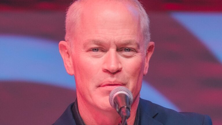 Neal McDonough fala ao microfone durante evento com iluminação vermelha e fundo desfocado ao palco