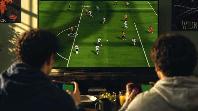 Pessoas jogam videogame de futebol na TV enquanto seguram smartphones em sala com pôsteres de séries da Netflix ao fundo