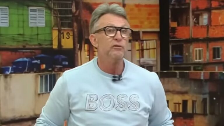 Neto de óculos e moletom azul durante comentário no Os Donos da Bola com imagem de comunidade ao fundo