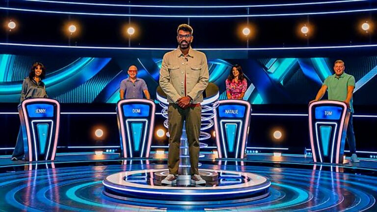 Apresentador Romesh Ranganathan no centro do palco com participantes posicionados em cabines do game show The Weakest Link