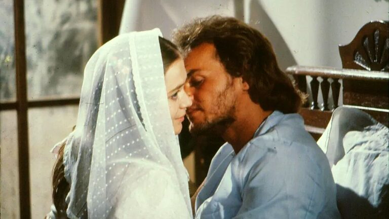 Casal troca momento de carinho à beira da cama em cena romântica de novela de época