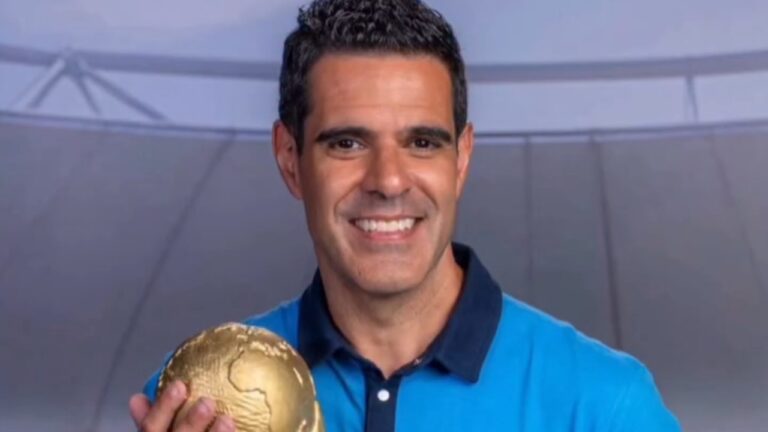 Paulo Andrade sorri segurando troféu em formato de globo usando camisa polo azul diante de cenário de estúdio esportivo