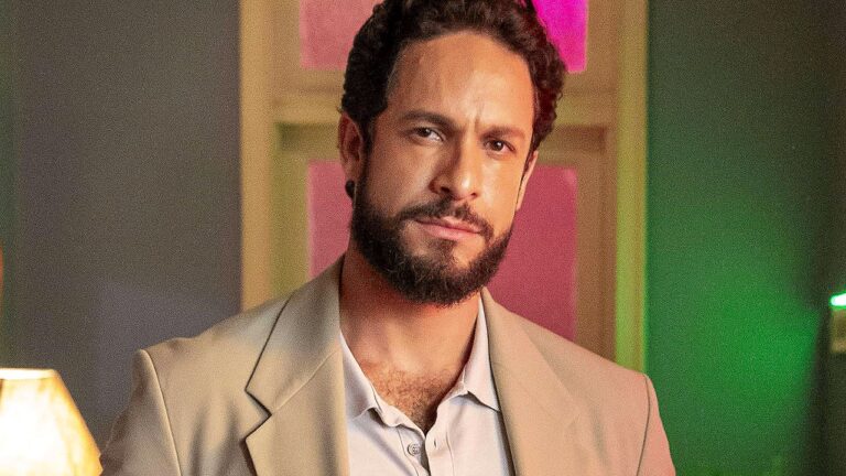 Ator de barba e blazer bege com expressão séria em cena de novela ambientada em casa noturna