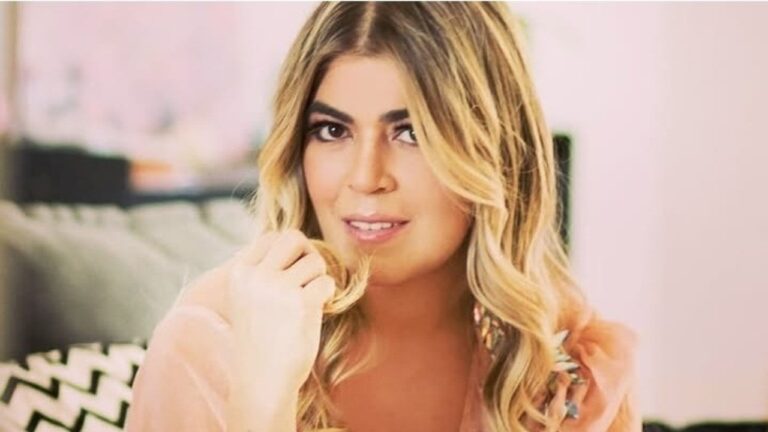 Raquel Pacheco com cabelo loiro ondulado segura mechas do cabelo e sorri levemente em ambiente interno com sofá ao fundo