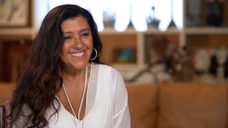 Regina Casé sorri durante entrevista em ambiente residencial usando blusa branca e acessórios prateados com estante desfocada ao fundo