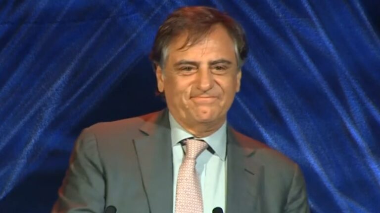 Ricardo Scalamandré de terno e gravata durante discurso em evento corporativo diante de painel azul iluminado
