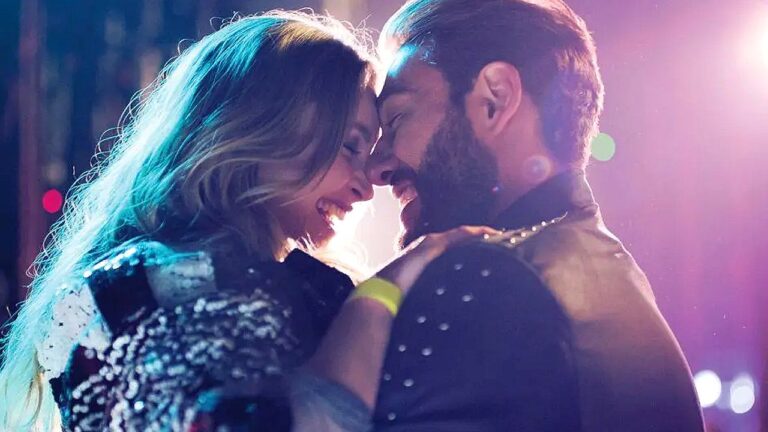 Casal se abraça e sorri durante momento romântico sob luzes coloridas em cena de filme ou série