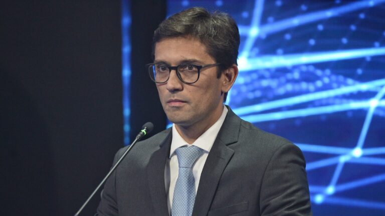 Rodolfo Schneider de terno escuro e gravata azul fala ao microfone diante de painel digital em debate da Band