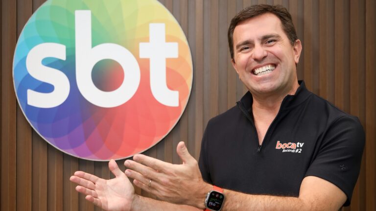 Rodrigo Bocardi sorri e gesticula diante do logotipo do SBT em foto corporativa na sede da emissora