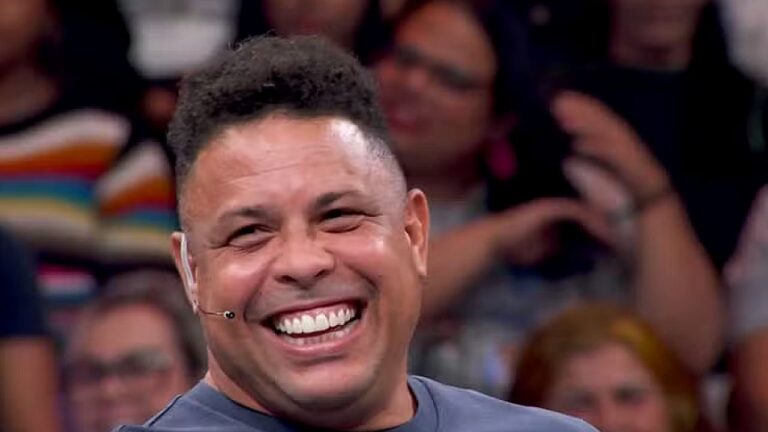 Homem sorrindo com microfone de cabeça durante participação em programa de auditório com plateia desfocada ao fundo