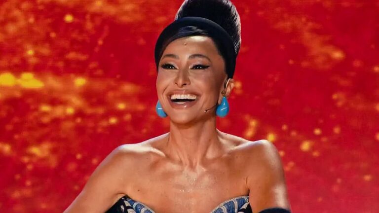Sabrina Sato sorrindo no palco de programa de TV com figurino inspirado em Carmen Miranda e fundo iluminado em vermelho