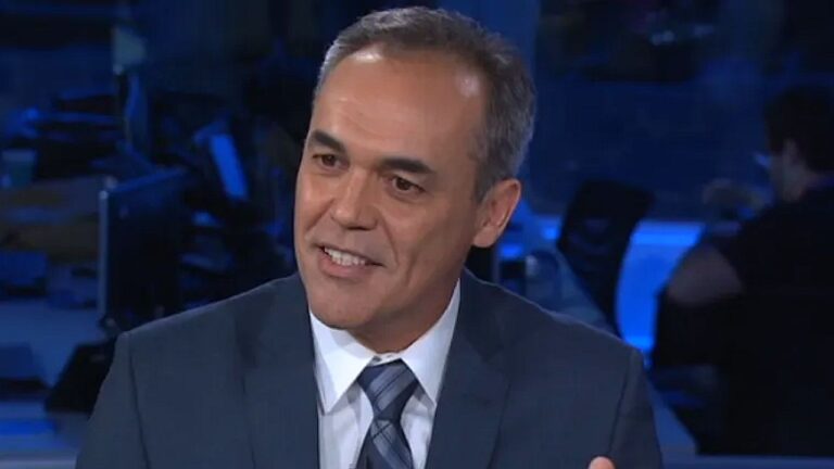 Jornalista de terno azul e gravata listrada fala durante entrevista em estúdio de telejornal com redação iluminada em tons de azul ao fundo