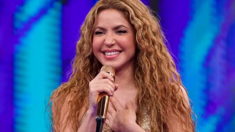 Shakira sorri enquanto segura microfone dourado e leva a mão ao peito durante apresentação em programa de televisão com cenário iluminado em tons de azul e roxo