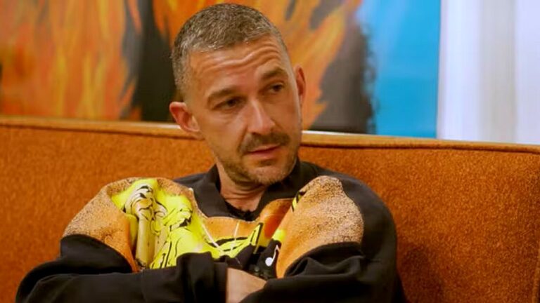 Shia LaBeouf sentado em sofá durante entrevista, usando moletom estampado em cenário de estúdio iluminado
